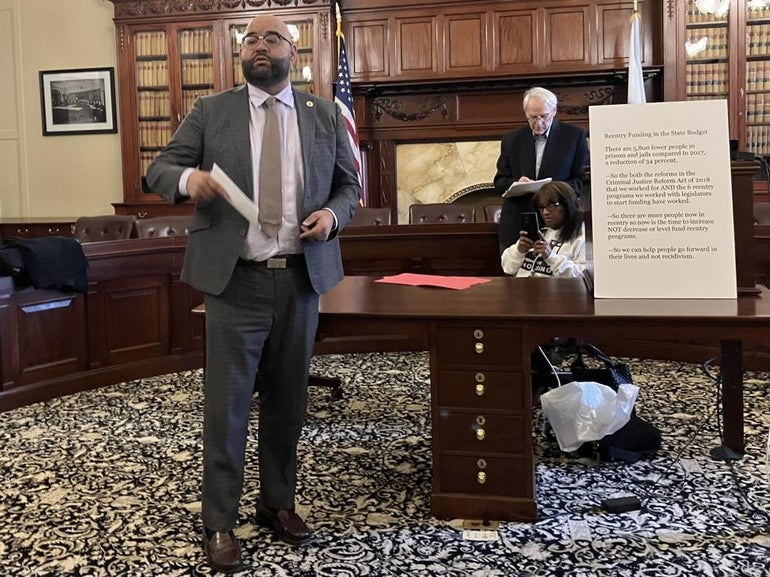 Three people in a carpeted room: one standing in front of a desk, one sitting behind a desk, and one standing behind the desk