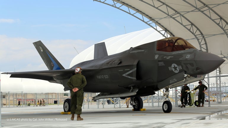 A Marine Corp member stands in front of a fighter jet preparing to taxi in a hanger