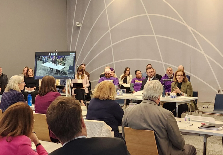 A woman speaks at a table with a microphone in front as a crowd looks on