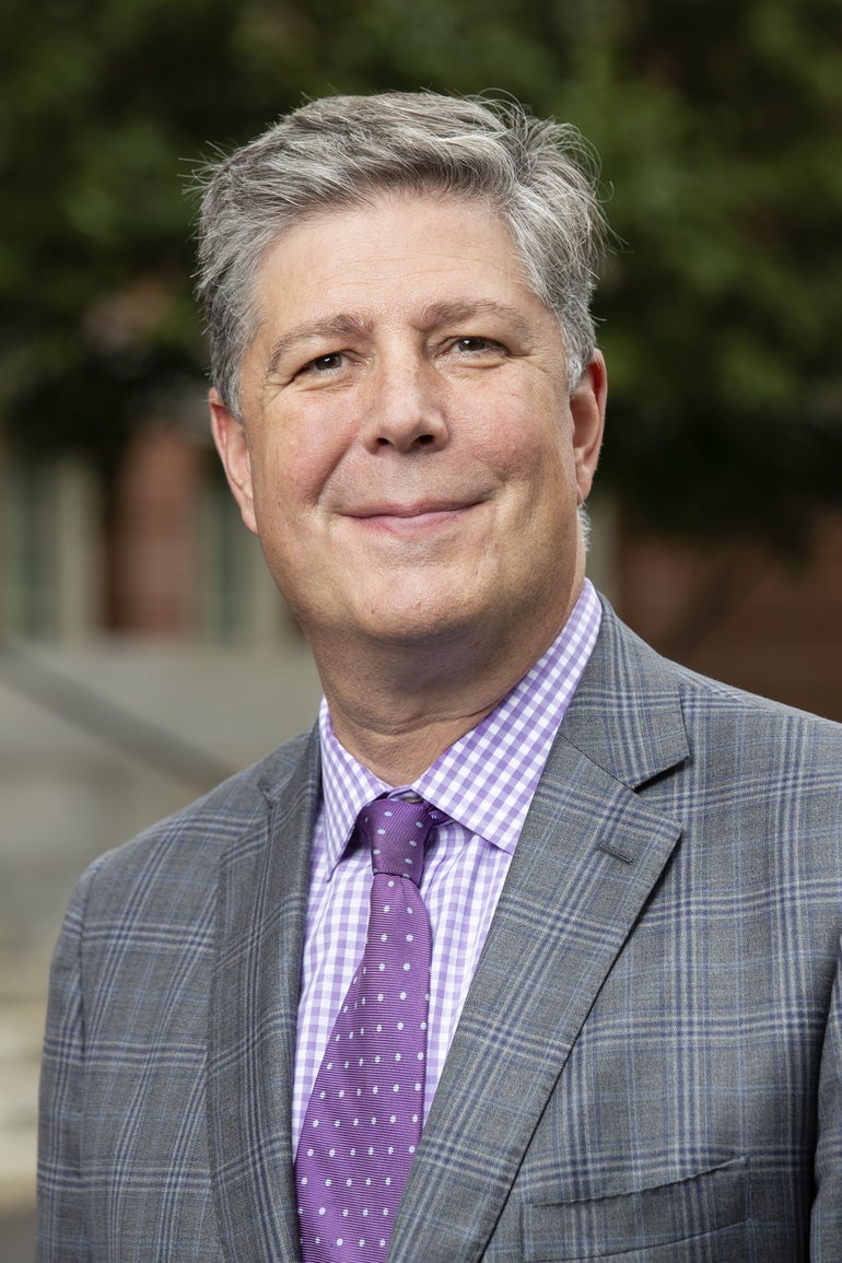 A man with gray hair wears a gray suit with a purple tie