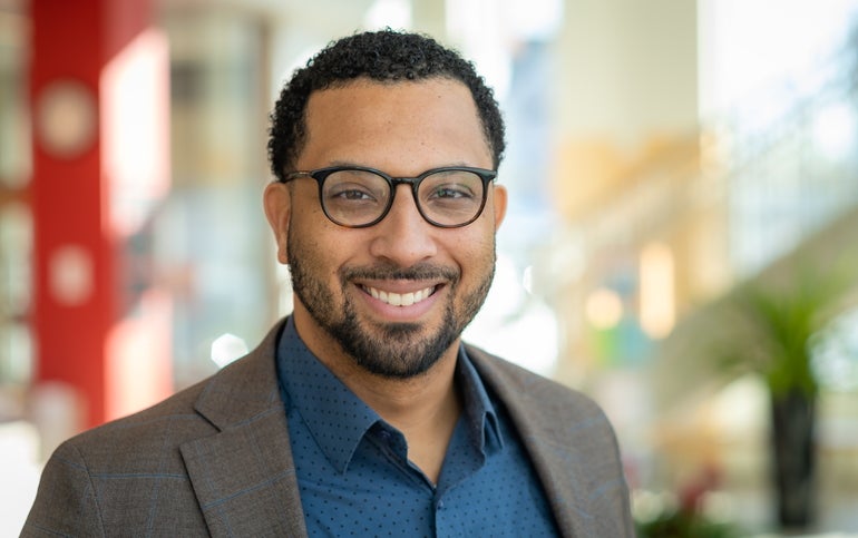 A man with dark hair, a beard, and black glasses wears a grey striped suit with a blue dotted button down.