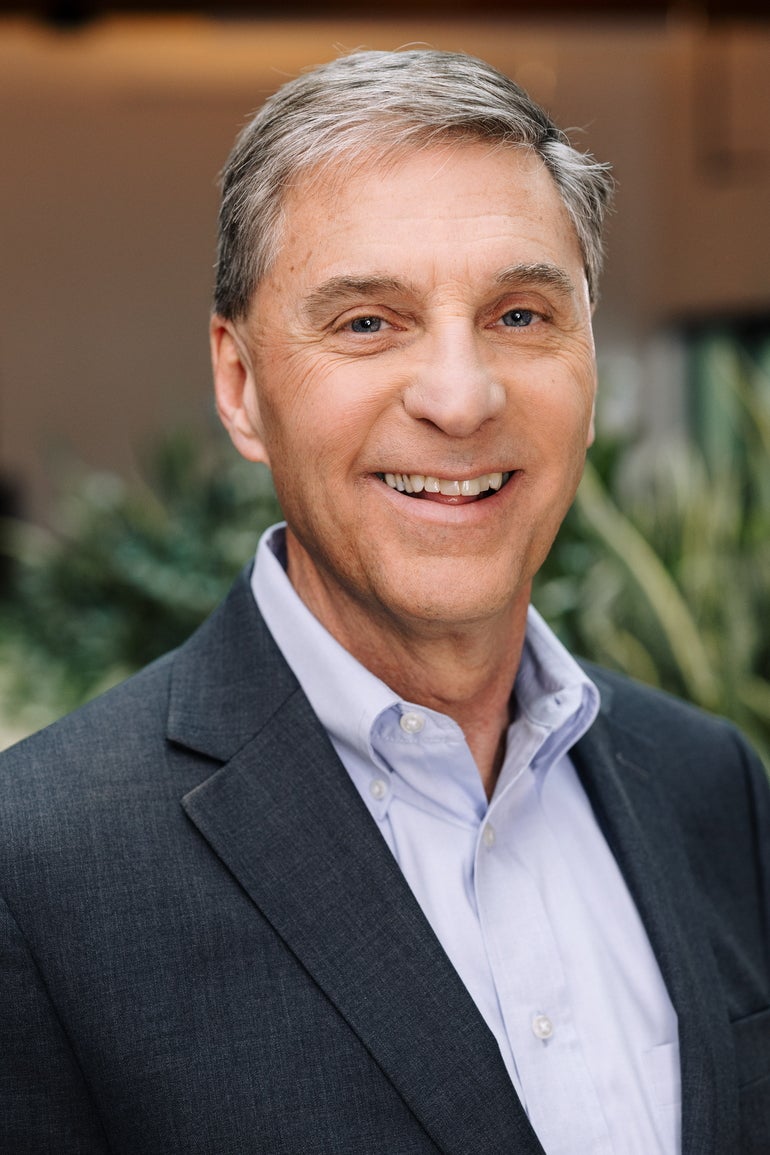 A man with salt-and-pepper hair wears a blue shirt and suit while smiling at the camera.