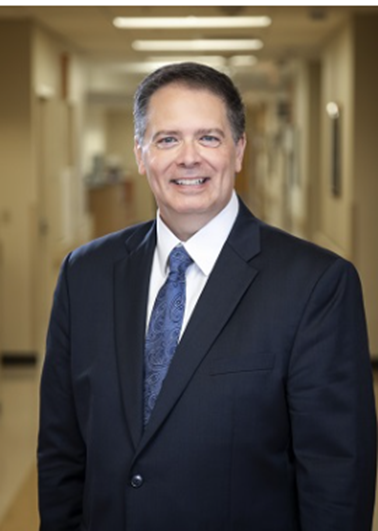 A man in a dark navy suit jacket, white shirt, and blue patterned tie