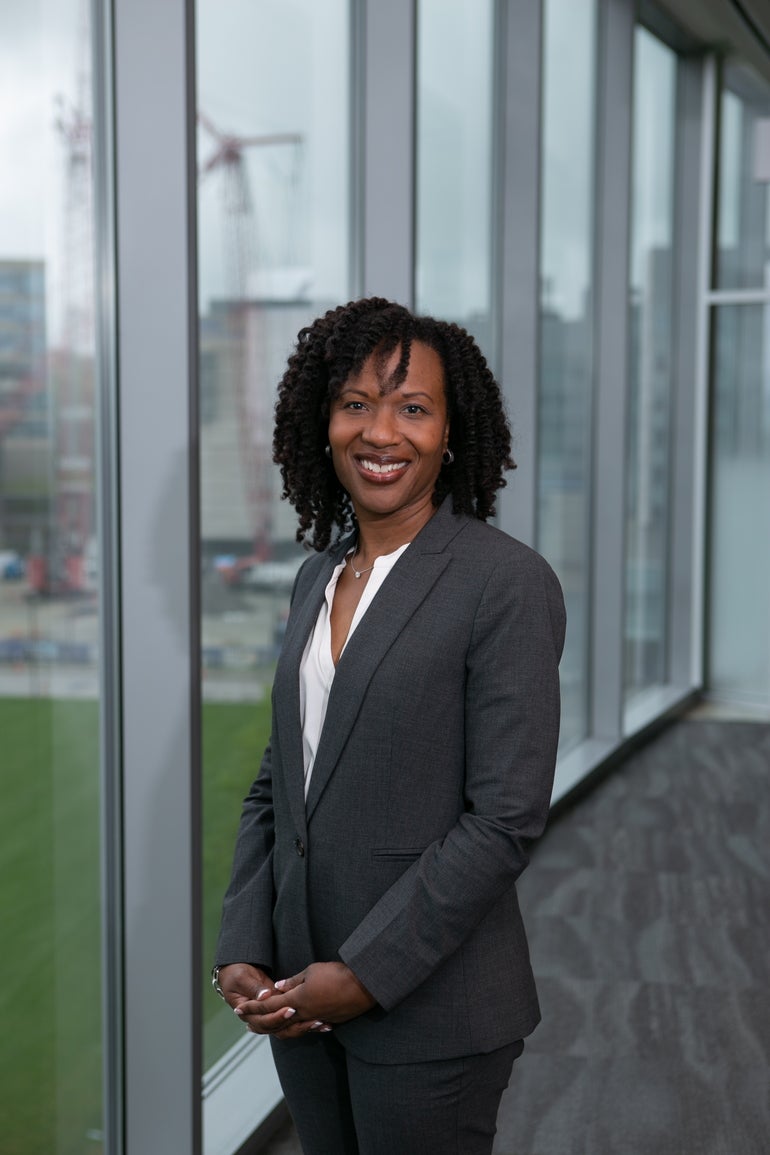 A woman with dark hair in a dark grey suit and white shirt stands in front of windows looking out to buildings and a grassy area.