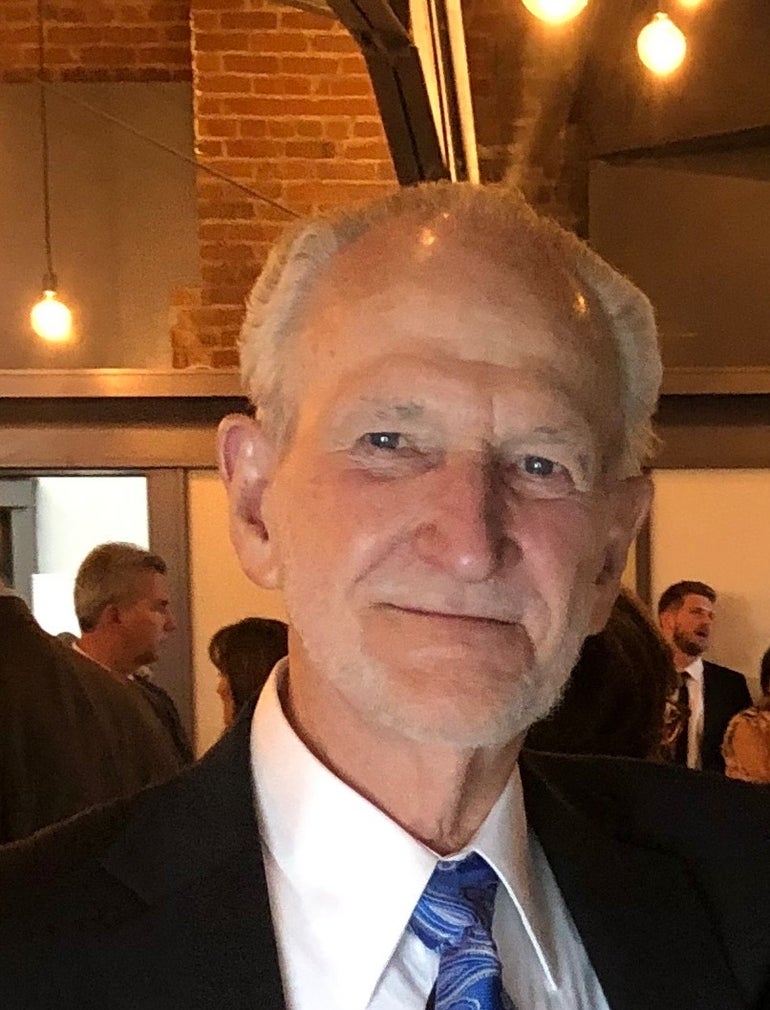 A man with white hair, a black suit jacket, white button down, and blue tie.