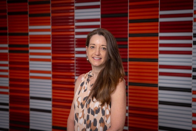 A woman with long brown hair stands in front of a colorful wall.