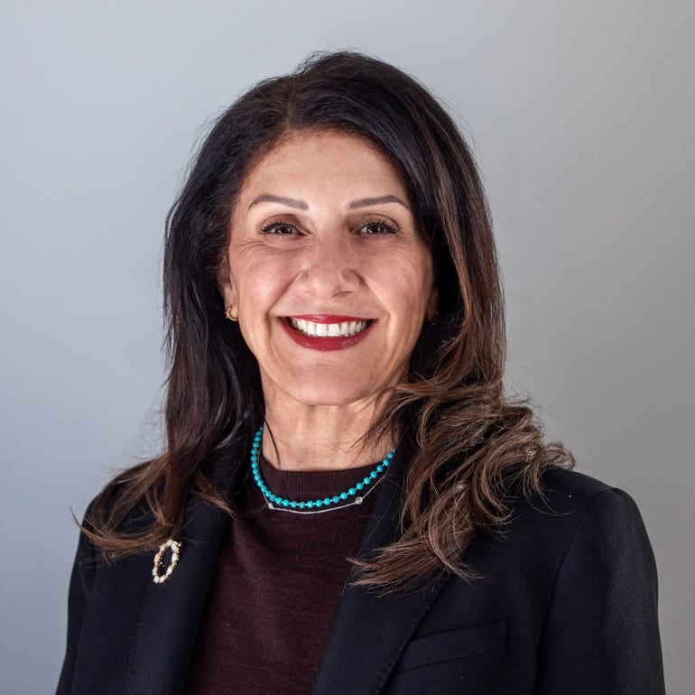 A woman with long, dark hair wears a brown sweater, black jacket, and blue necklace.