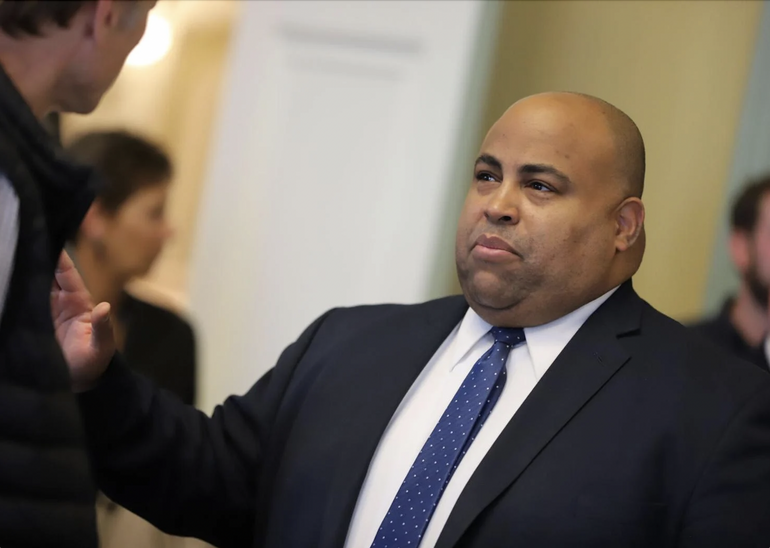 A man in a black suit jacket, white shirt, and blue tie engages in an exchange with a man in a black vest.