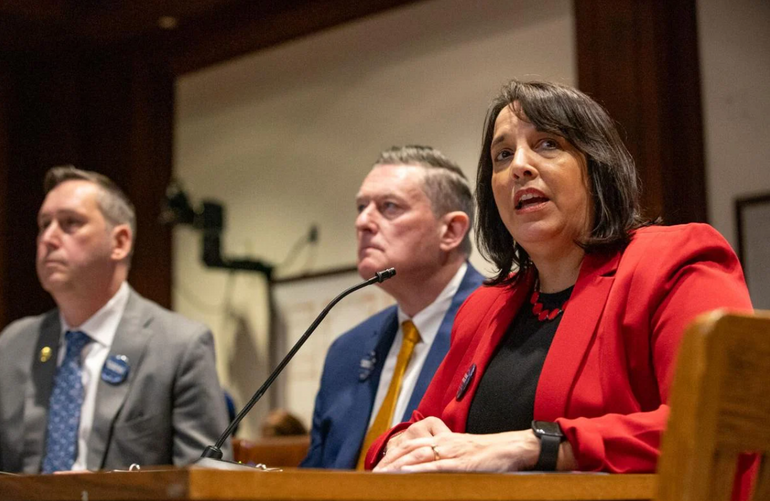 A woman speaks into a microphone at a table during a hearing