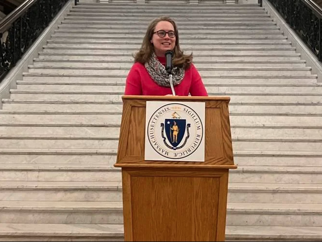 Rep. Christine Barber wears a pink shirt and patterned scarf standing behind a podium in front of a wide staircase