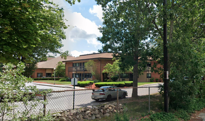 Two brick residential buildings, one with a balcony, peeking out behind green trees