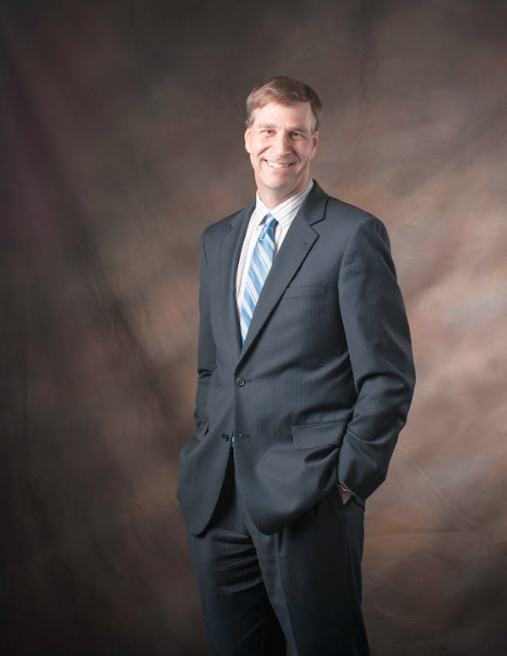 A man with brown hair standing in a dark suit, striped button down, and blue diagonally striped tie.