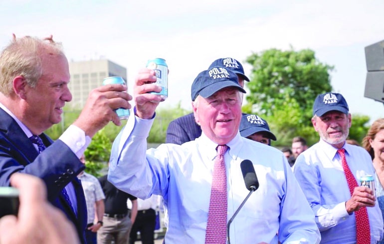 Men in light blue shirts, red ties, and dark blue baseball caps hold up blue cans of seltzer outside.