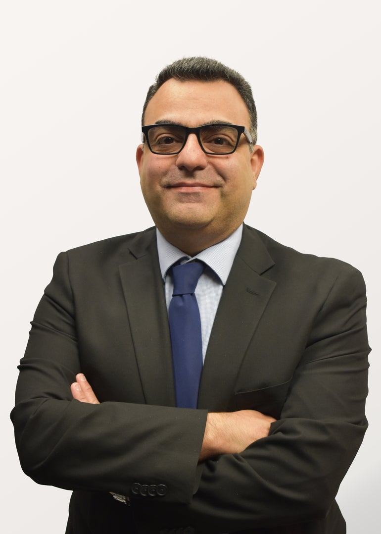A man with glasses, a black suit jacket, light blue button down, and dark blue tie stands with crossed arms.