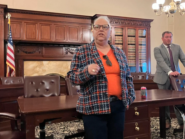 A woman in an orange shirt and black and orange printed jacket stands in front of a large brown desk with brown leather chairs behind.