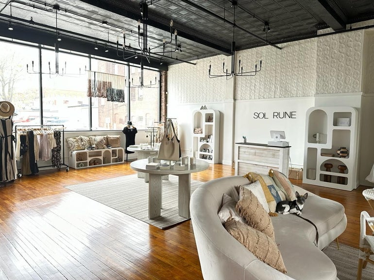 An expansive retail space with various clothes items on a rack in the back of the space near windows. A point of sale system and desk sit to the right. A couch with a small dog on in sits in front. 