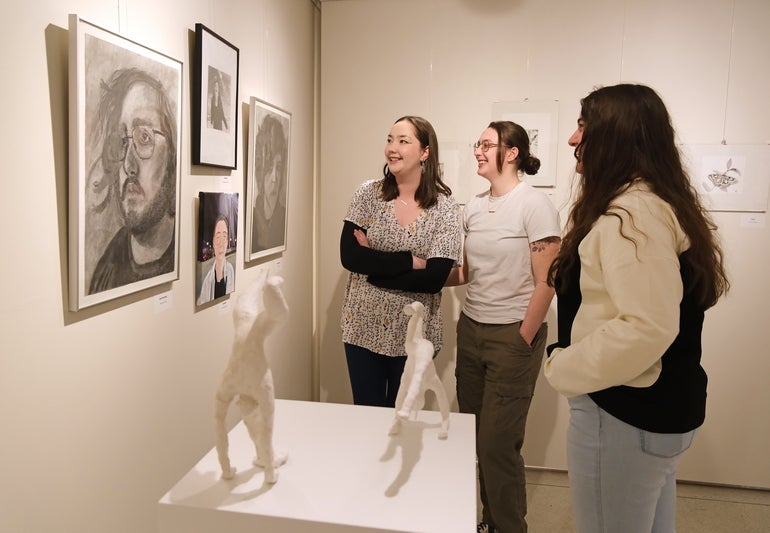 Three people look at photos and paintings of people in a white-walled gallery.