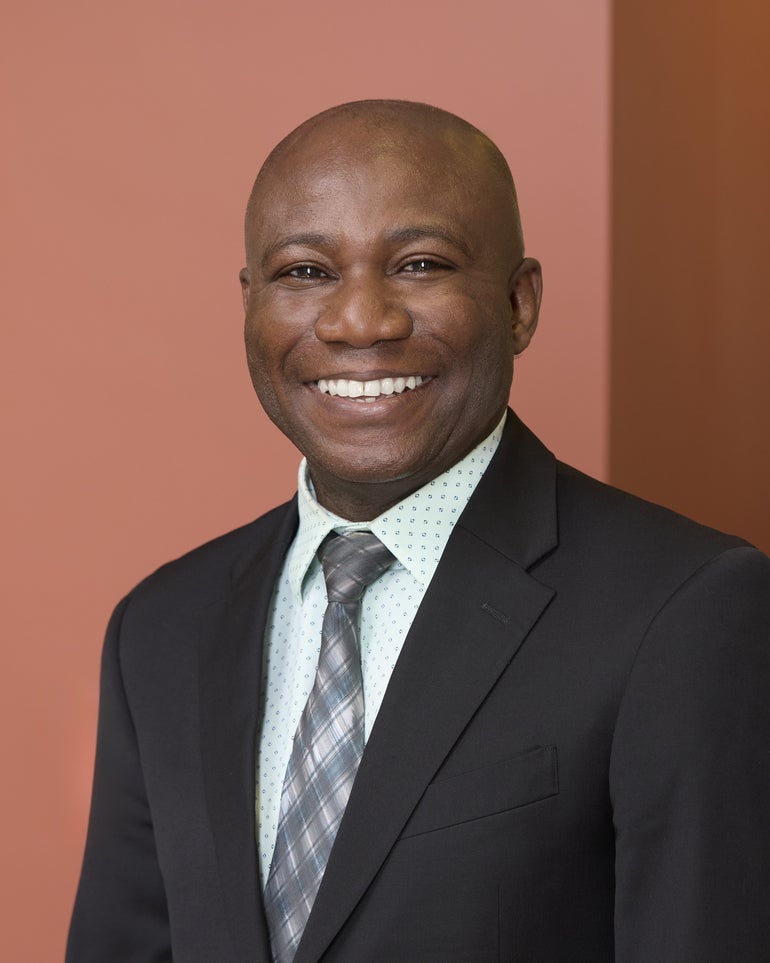 A man wears a black suit jacket, white printed button down shirt, and a blue, grey, and white patterned tie.