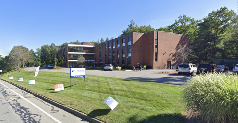 A brick building sits behind a parking lot with a green lawn separating it from the street.