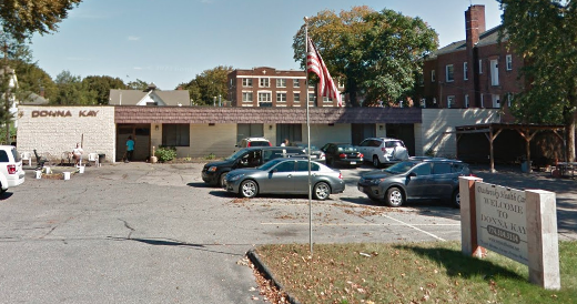 A brick building sits behind a cracked paved parking lot with an American flag flying in front.
