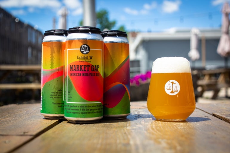 A pour of beer next to a four pack, on a picnic table on a brewery patio