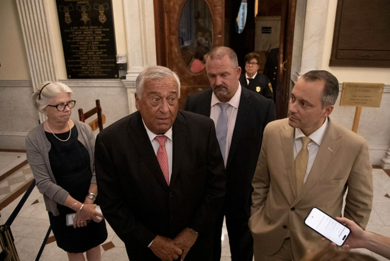 A man with white hair, a black suit jacket, white button down, and red tie speaks to a reporter off camera who is holding a phone up in frame to record him.