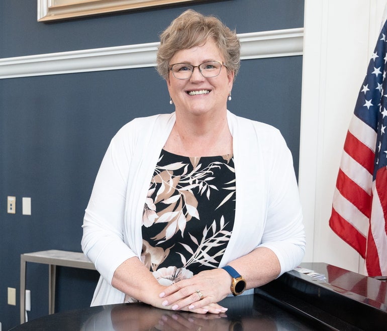 A woman with short grey hair wears a white cardigan and a black/beige/white floral print blouse. She stand with her hands together while leaning against a piano with an American flag on the right.