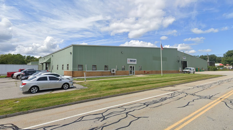 A large green industrial building sits behind a road with cars parked next to it