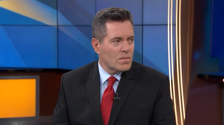 A man wears a black suit jacket, light blue button down, and red tie.