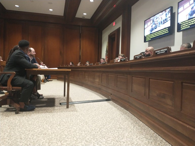 Three people sit at a table testifying to lawmakers in a hearing room