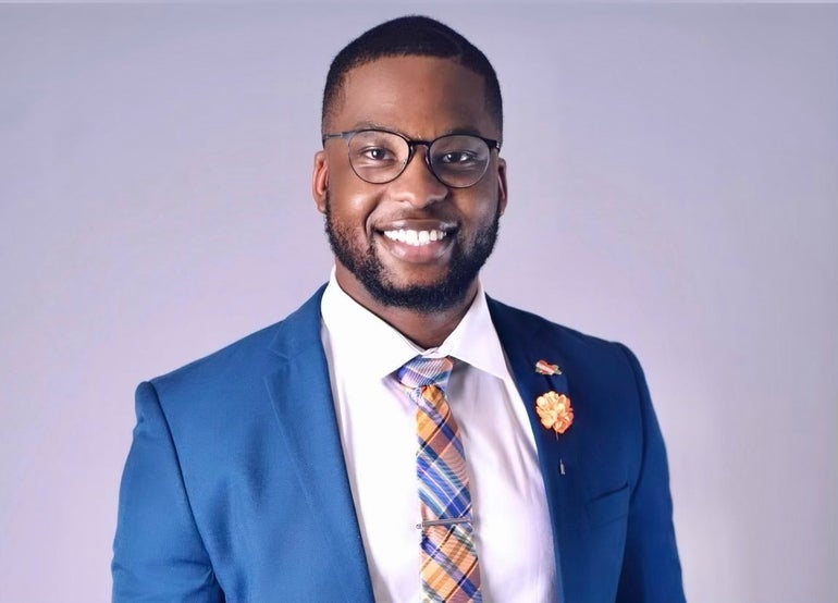 A man with black glasses wears a blue suit jacket, white button down, and blue, yellow, and white plaid tie