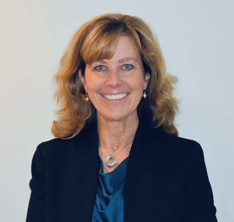 A woman with shoulder length blonde hair and bangs wears a black suit jacket and a blue silk blouse