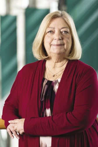 A woman with chin length blonde hair wears a red sweater and black, white, and purple patterned blouse.