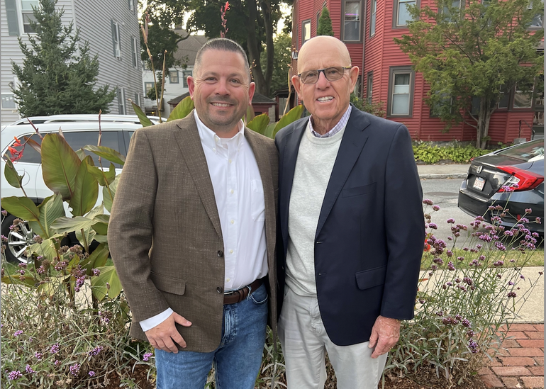 Two men stand next to each other in front of purple flowers that are planted in front of a house-lined street. The man on the left wears a brown suit jacket, white button down, and jeans while the man on the right wears a dark blue suit jacket, grey sweater, and white pants.