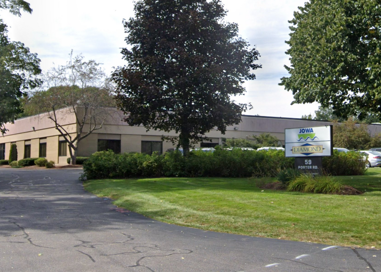 A one-story industrial building behind a lawn, trees, and a bush