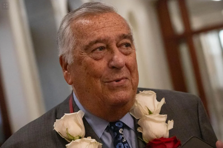 A man with grey hair wears a grey suit jacket, blue button down, blue and white printed tie, and flowered necklace