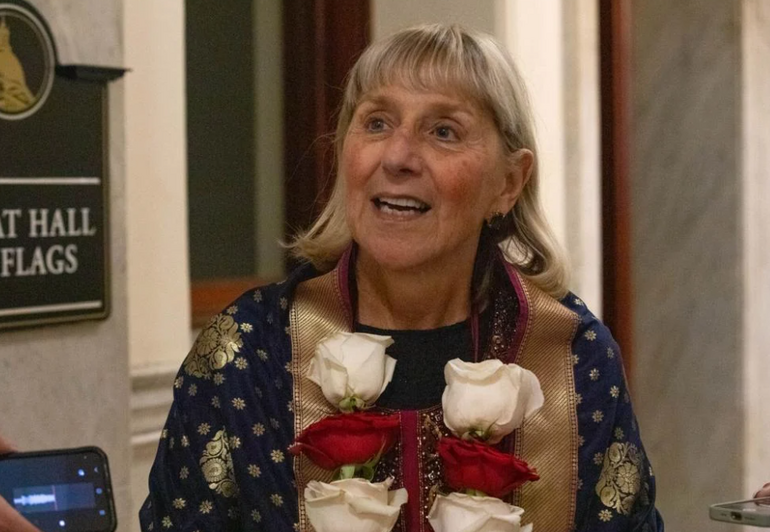 A woman with shoulder length grey hair and bangs wears a flower necklace while speaking to a reporter off camera holding a phone recording her on the bottom left of the photo.