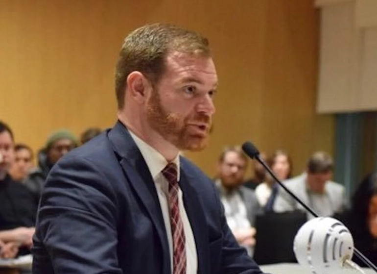 A man with red hair and a red beard wears a dark blue suit jacket, a white button down, and a red patterned tie while speaking into a microphone