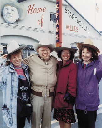 A man and three women stand with their arms around each other all wearing cowboy hats outside in front of a store