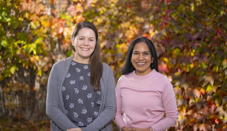 Two women stand side by side in front of colorful tree leaves. The woman on the left has long brown hair and wears a grey cardigan and grey floral-patterned shirt. The woman on the right is shorter with long black hair and wears a light pink sweater.