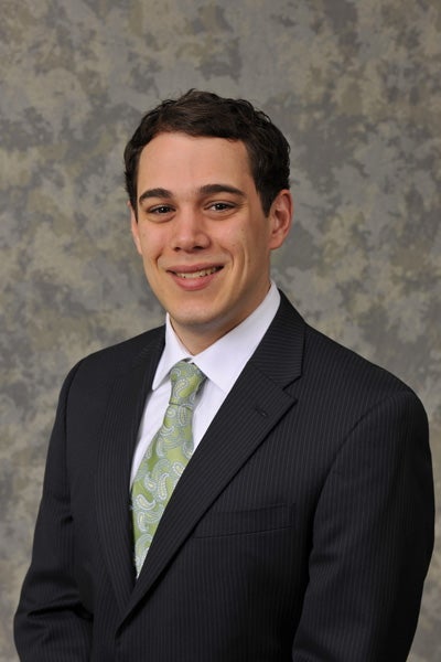 A headshot of a man with short dark brown hair, a black suit jacket, white button down, and light green paisley tie