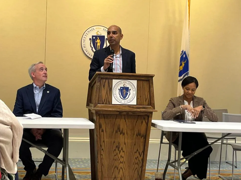 A man wearing a black suit jacket and pink and blue plaid button down stands at a podium with a man wearing a dark blue suit and light blue button down sitting at a table to his the and a woman wearing a tan suit jacket and white top sitting at a table to the right.