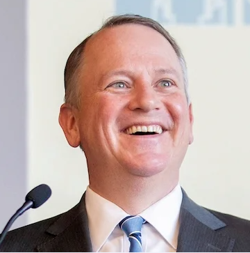 A man with grey hair wears a black suit jacket, white button down, and a blue striped tie.