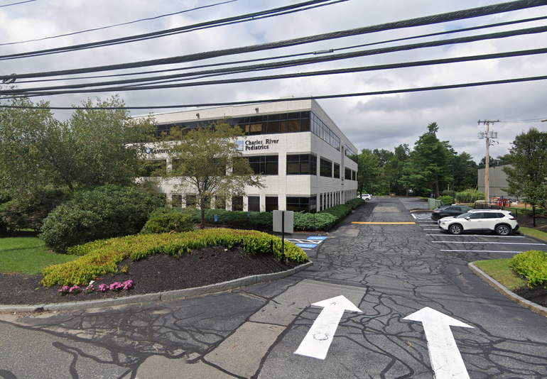 A white three-story office building sits behind some trees, viewed from the entrance to its parking lot