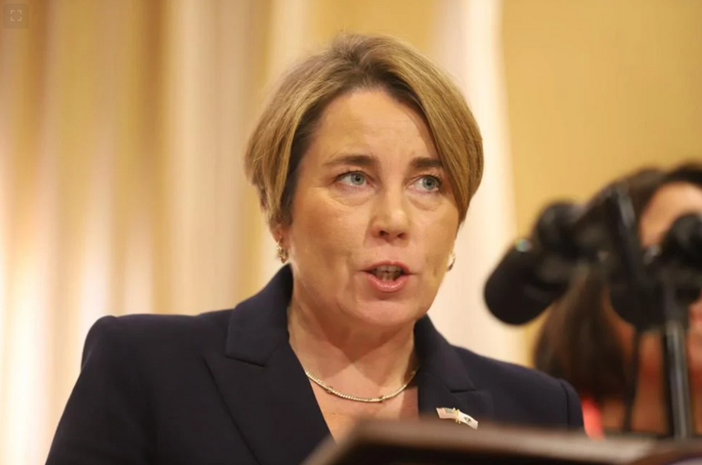 A woman with blonde hair wearing a a dark blue suit jacket with an American flag pin on it stands speaking at a podium