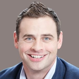 A man with light brown hair smiles for the camera wearing a dark blue suit jacket and white button down