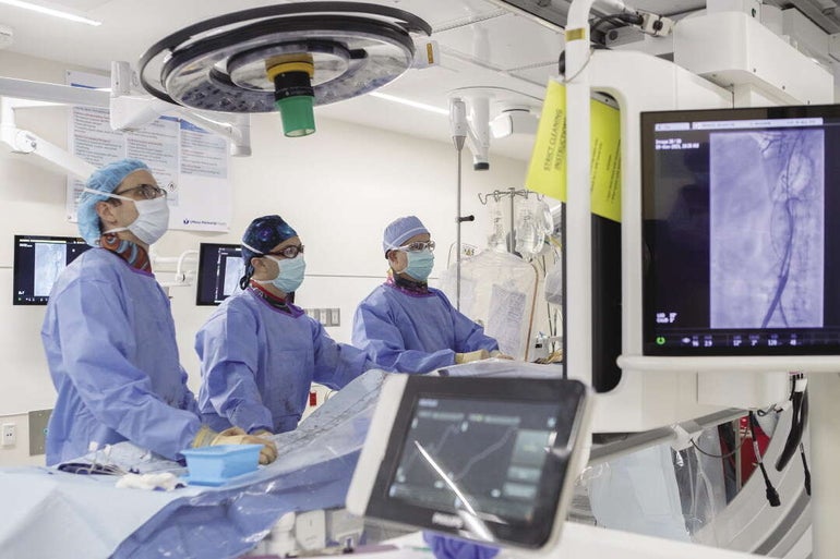 Three doctors in light blue surgical gowns and masks stand in an operating room looking at a screen.