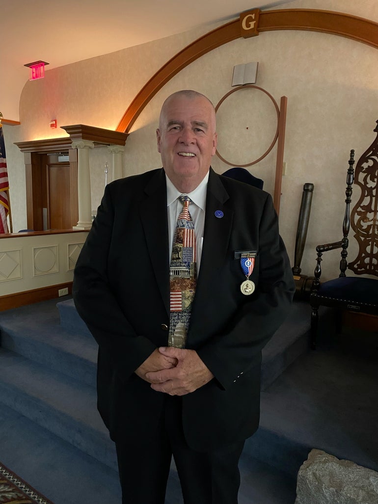 A man stands with his hands together in a black suit and patterned tie