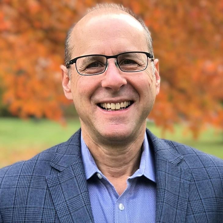 A man wears black glasses, a blue plaid suit jacket, and light blue button down while standing in front of colorful trees.