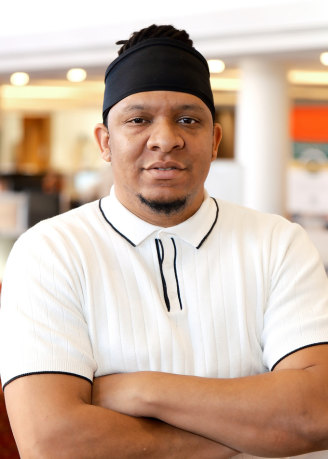 A man wearing a black head wrap wears a white polo with black lining with his arms crossed in front of him.